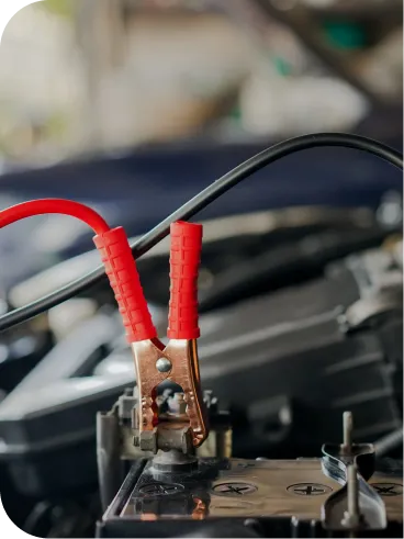 A close-up of red jumper cables attached to a car battery terminal under the hood, with engine components visible in the blurred background.