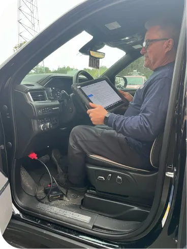 A man sits in the driver’s seat of a car, holding a tablet connected via cable to the vehicle, likely conducting a diagnostic or inspection. He wears sunglasses and dark clothing. The car door is open.