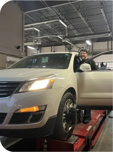 A person wearing a cap stands by the open driver’s door of a white SUV on a car lift in an auto repair shop, smiling at the camera. The vehicle’s headlights are on and equipment is attached to the front wheel.