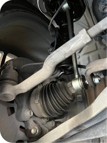 Close-up view of a cars suspension system, showing components like the shock absorber, control arm, tie rod, and CV axle with a rubber boot, all covered in a layer of dust.