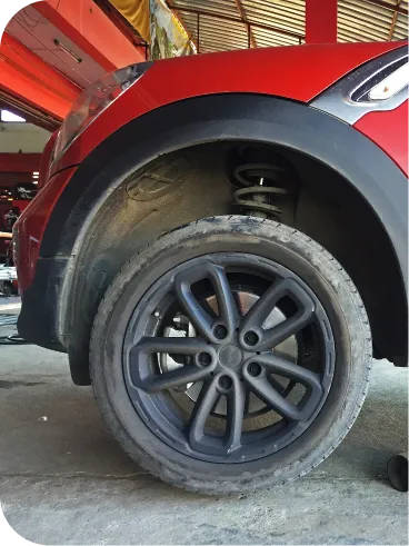 Close-up of the rear wheel and tire of a red car, showing the suspension system and wheel well. The car is parked inside a garage with a concrete floor and metal roof visible in the background.