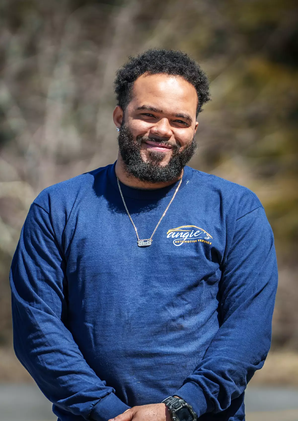 Portrait of Alex, Technician at Angie's Automotive Service, wearing a blue long-sleeve company shirt with logo on the chest, a pendant necklace, and a watch on the left wrist, standing outdoors with softly blurred trees in the background.