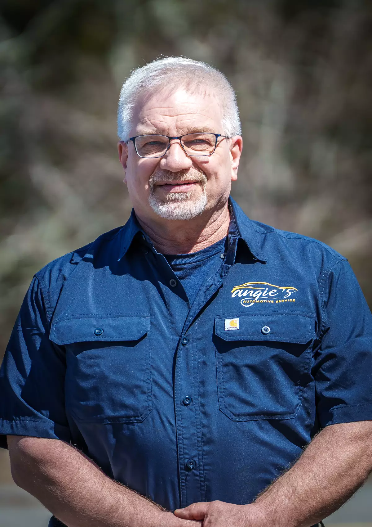 A middle-aged man with short gray hair, glasses, and a beard stands outdoors, wearing a blue button-up shirt with an embroidered logo that reads Angies Custom Design. The background is blurred.