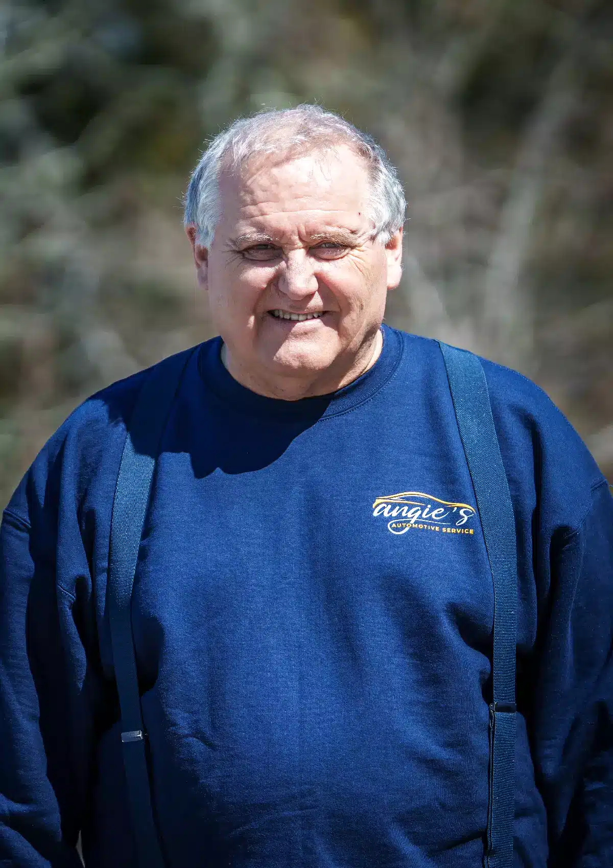 Portrait of Gary, owner of Angie's Automotive Service, wearing a dark blue shop sweatshirt and matching suspenders, standing outdoors with softly blurred trees in the background.