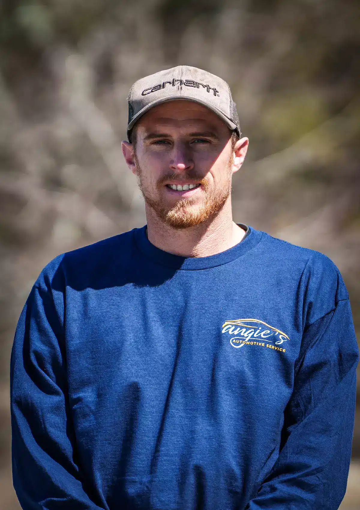 Portrait of James, Technician at Angie's Automotive Service, wearing a blue long-sleeve shop shirt with company branding and a beige Carhartt cap, standing outdoors with softly blurred greenery in the background.