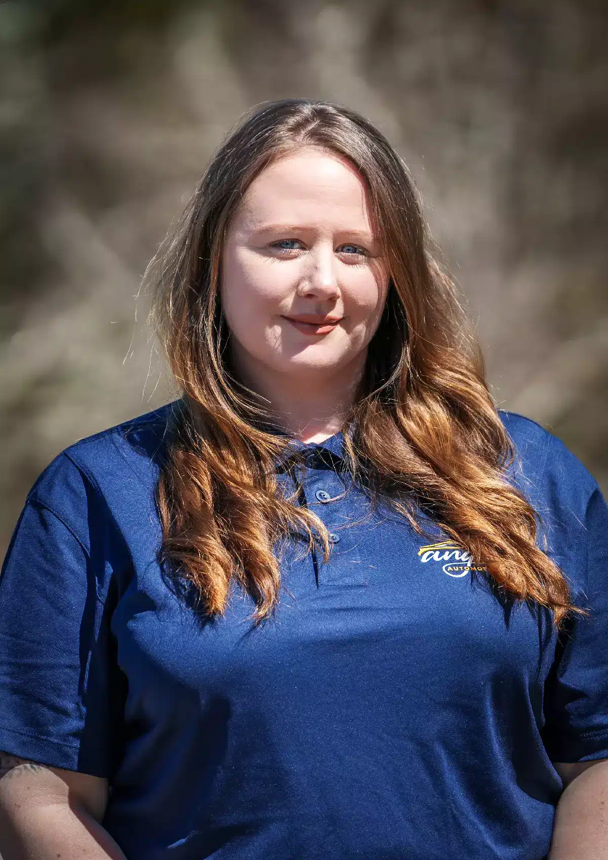 Portrait of Nicolette, Sales Administrator and Marketing Specialist at Angie's Automotive Service, wearing a blue polo shirt with the Tate Automotive logo, standing outdoors with a softly blurred background. She has long, wavy hair and a poised, friendly expression.