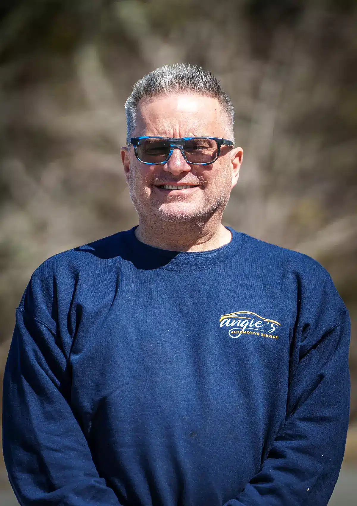 Portrait of Rob, Diagnostician and ADAS Calibrator at Angie's Automotive Service, wearing a navy blue company sweatshirt, standing outdoors with a background of softly blurred trees.