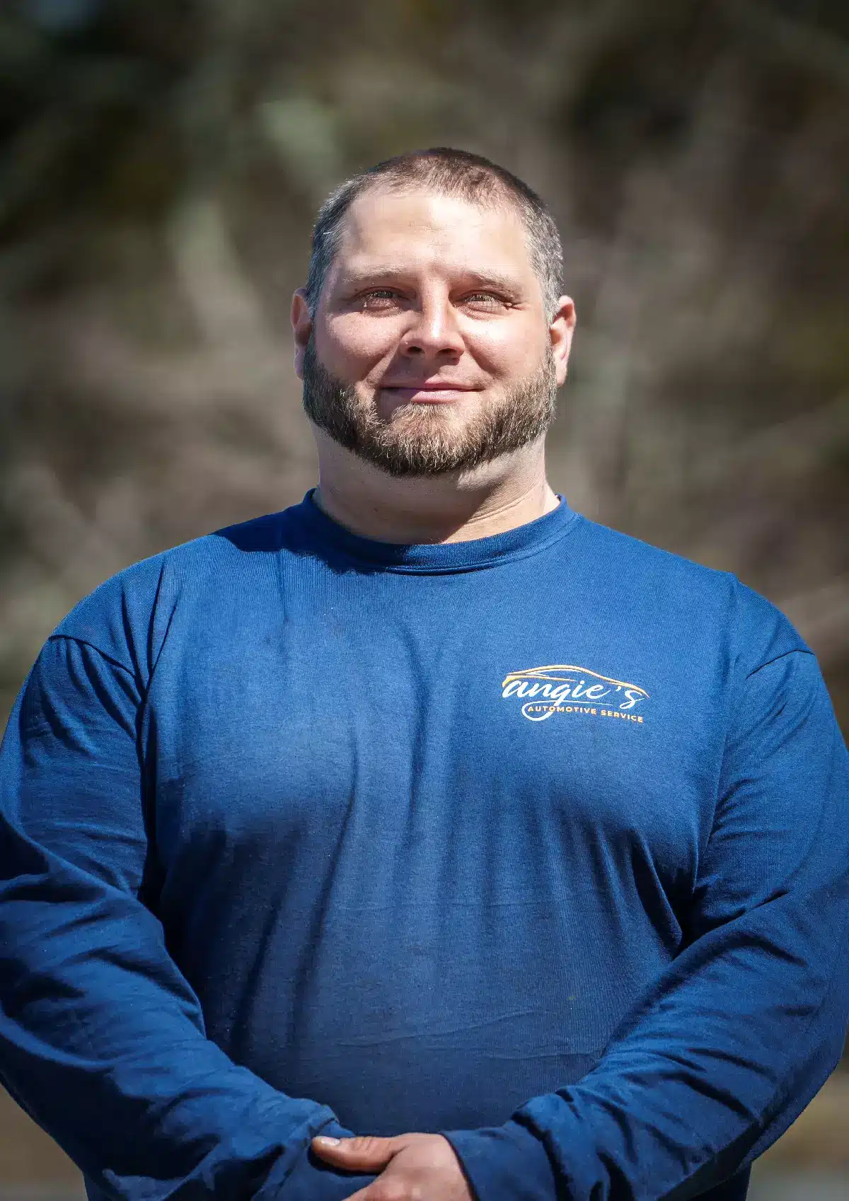 Portrait of Tim, Technician at Angie's Automotive Service, wearing a blue long-sleeve company shirt with logo on the chest, standing outdoors against a softly blurred natural background.