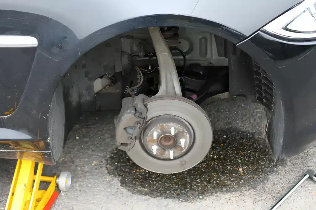 Car lifted on a jack stand with the wheel removed, exposing the brake rotor, caliper, and suspension components, with fluid leaking onto the ground.