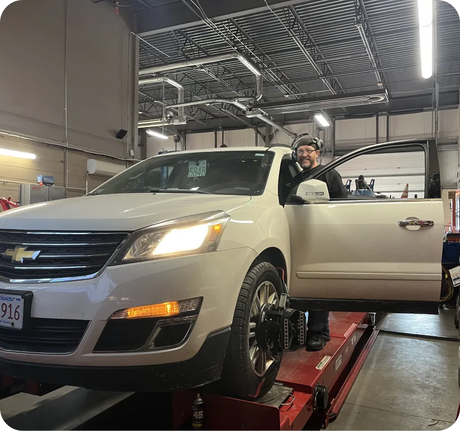 A mechanic stands by the open driver’s door of a white Chevrolet SUV on a lift inside a well-lit automotive workshop, smiling at the camera. The vehicles front wheels are mounted with alignment equipment.