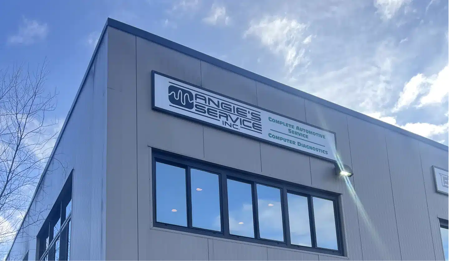 A modern gray building with large reflective windows and a sign that reads Angies Service Inc. Complete Automotive Service, Computer Diagnostics. The sky is partly cloudy.