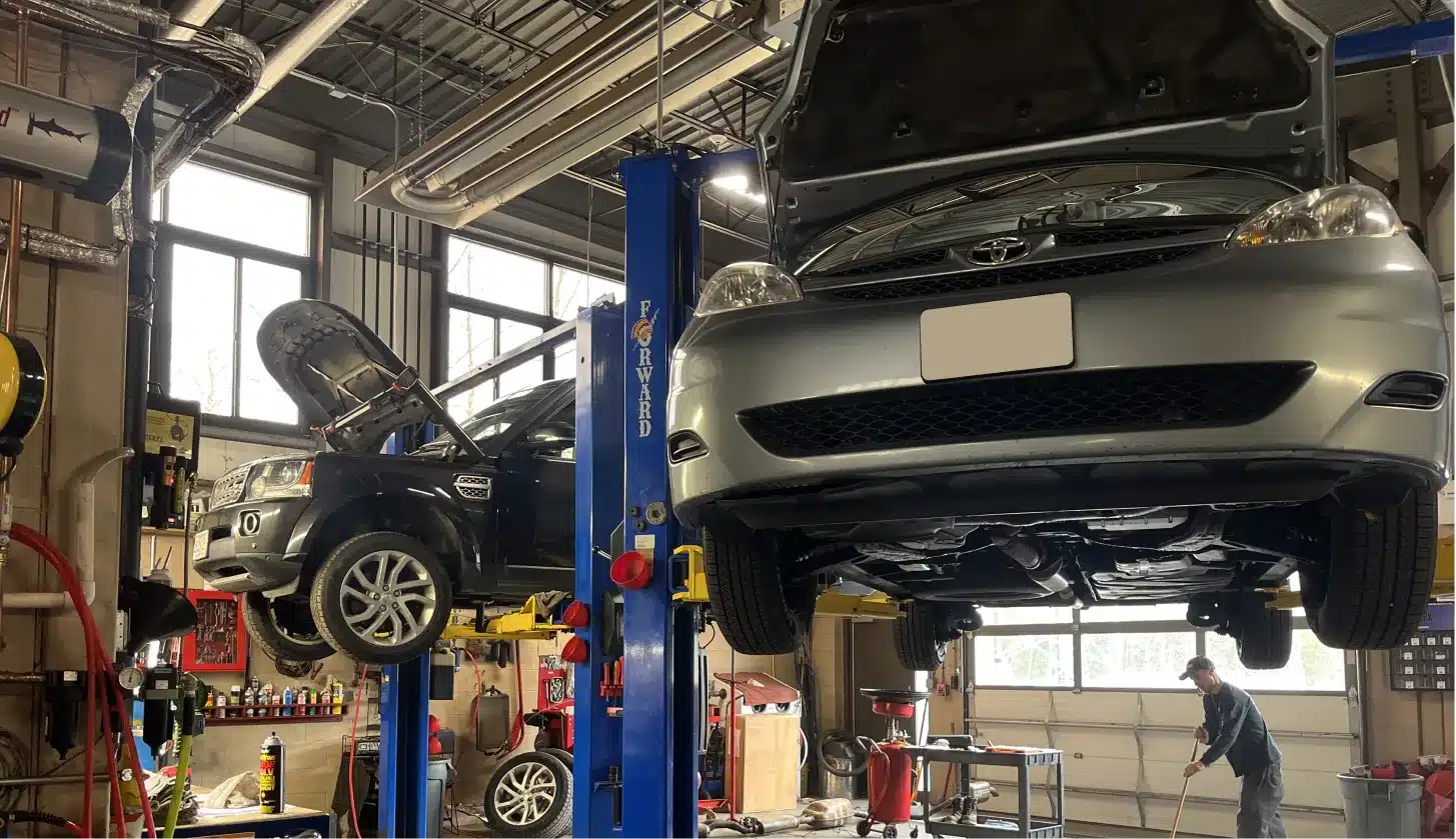 Two cars are lifted on hydraulic platforms in a bright auto repair shop. Mechanics tools and equipment are visible, and a person works near the rear of the garage. Both cars have their hoods open.