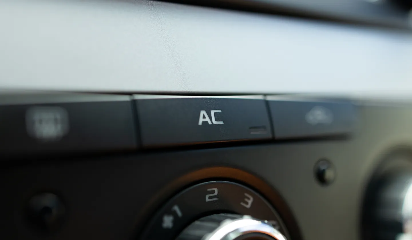 Close-up of a car dashboard showing the AC button for air conditioning, surrounded by other control buttons and part of a temperature dial with numbers visible.