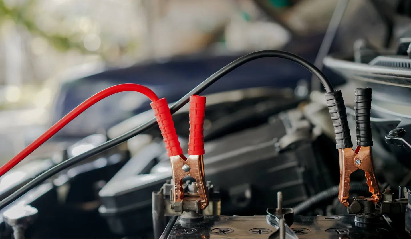 Close-up of jumper cables attached to a car battery, with one red and one black clamp connected to the battery terminals under an open car hood.