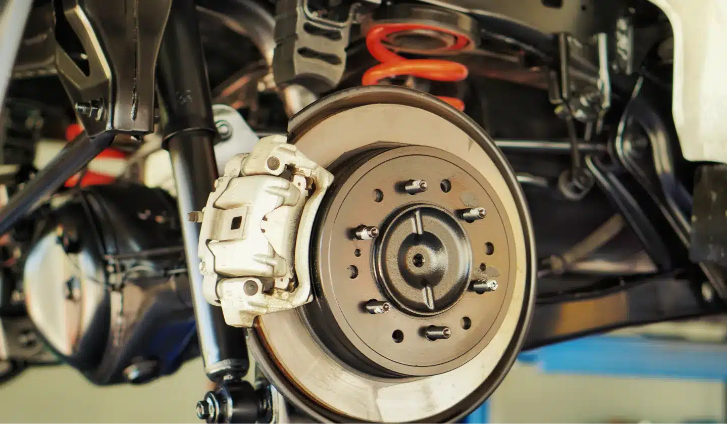 Close-up view of a car’s suspension system showing a disc brake, caliper, red coil spring, and various metal components, all part of the vehicle’s undercarriage, in a well-lit garage or workshop.