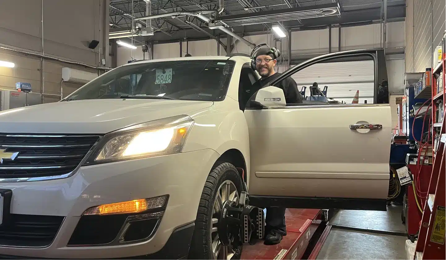 A mechanic stands beside an open driver’s door of a white Chevrolet SUV in an auto repair shop, with the vehicle positioned on a lift for alignment and diagnostic equipment attached to the front wheel.