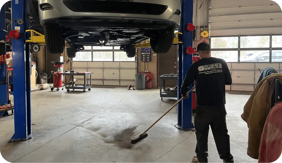 A mechanic sweeps the floor in a garage, with a car lifted on a hydraulic lift and tools visible in the background.