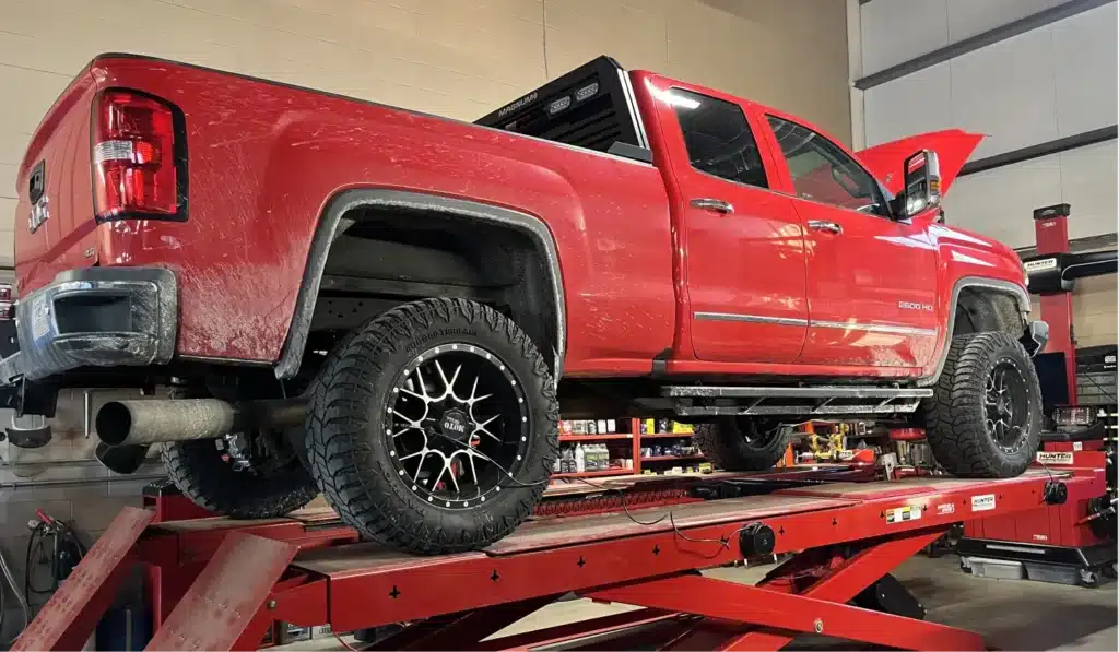 A red pickup truck is elevated on a hydraulic lift inside an auto repair shop. The truck’s hood is open, and large black wheels with chrome accents are visible. Tools and equipment are seen in the background.