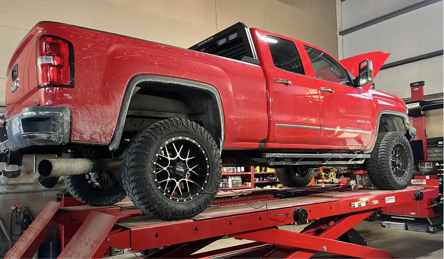 A red pickup truck is elevated on a hydraulic lift inside an auto repair shop. The truck’s hood is open, and large black wheels with chrome accents are visible. Tools and equipment are seen in the background.
