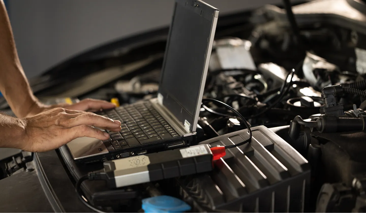A person uses a laptop connected to a device on a car engine, likely performing diagnostics or tuning in an automotive repair or maintenance setting.