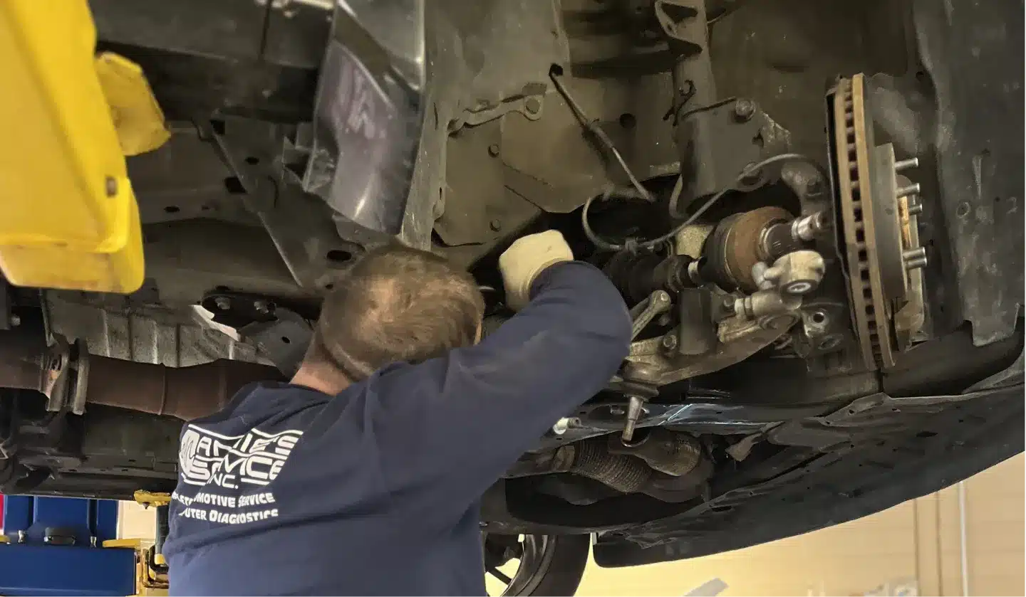 A mechanic wearing gloves works underneath a raised car, focusing on the vehicle’s suspension and brake components in an auto repair shop.