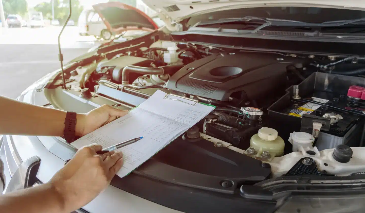 A person holds a clipboard and pen, checking a checklist next to an open car engine bay, suggesting an automotive inspection or maintenance process.