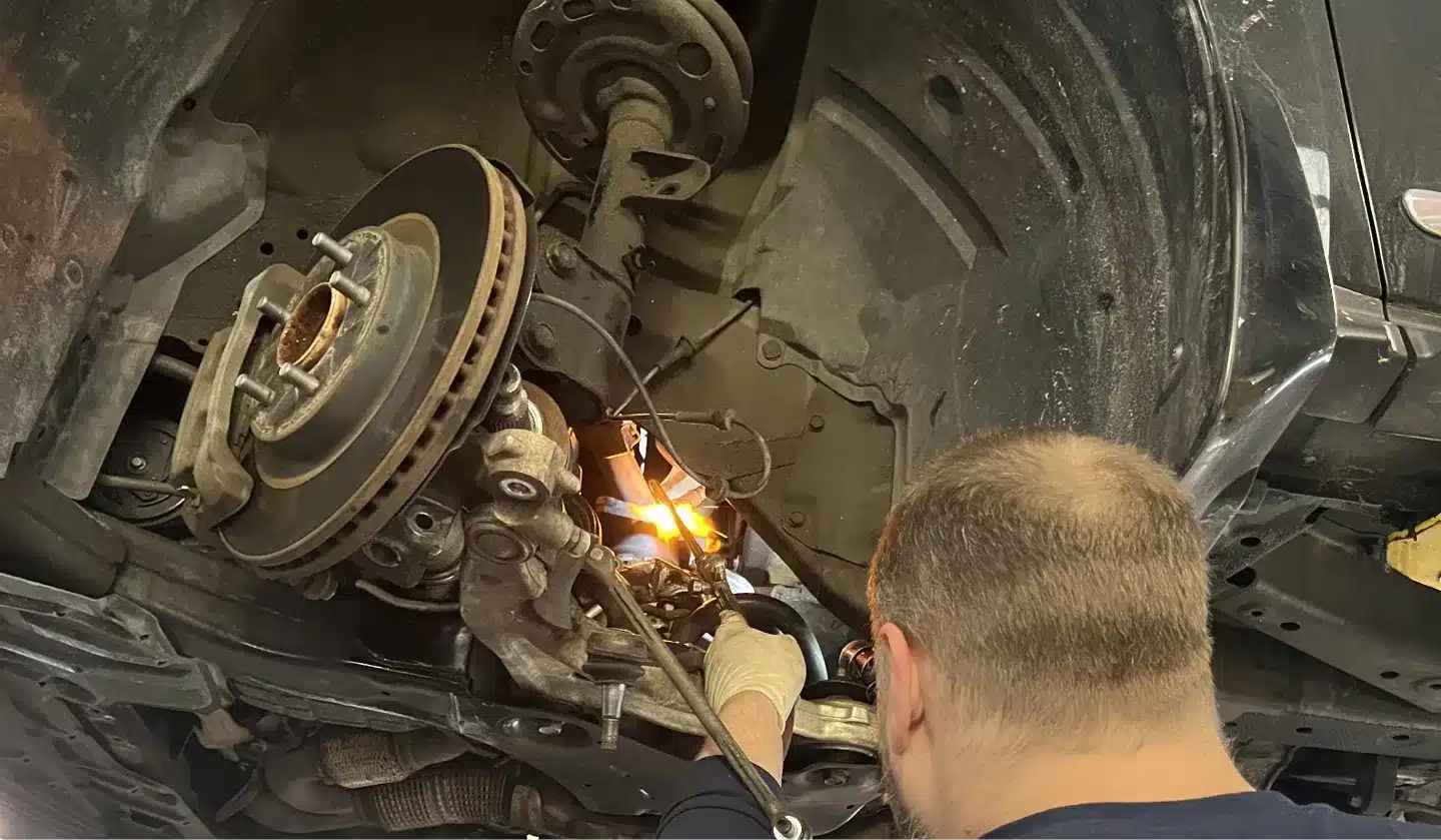 A mechanic works underneath a raised vehicle, focusing on the suspension and brake system, with sparks flying from welding or cutting equipment nearby.