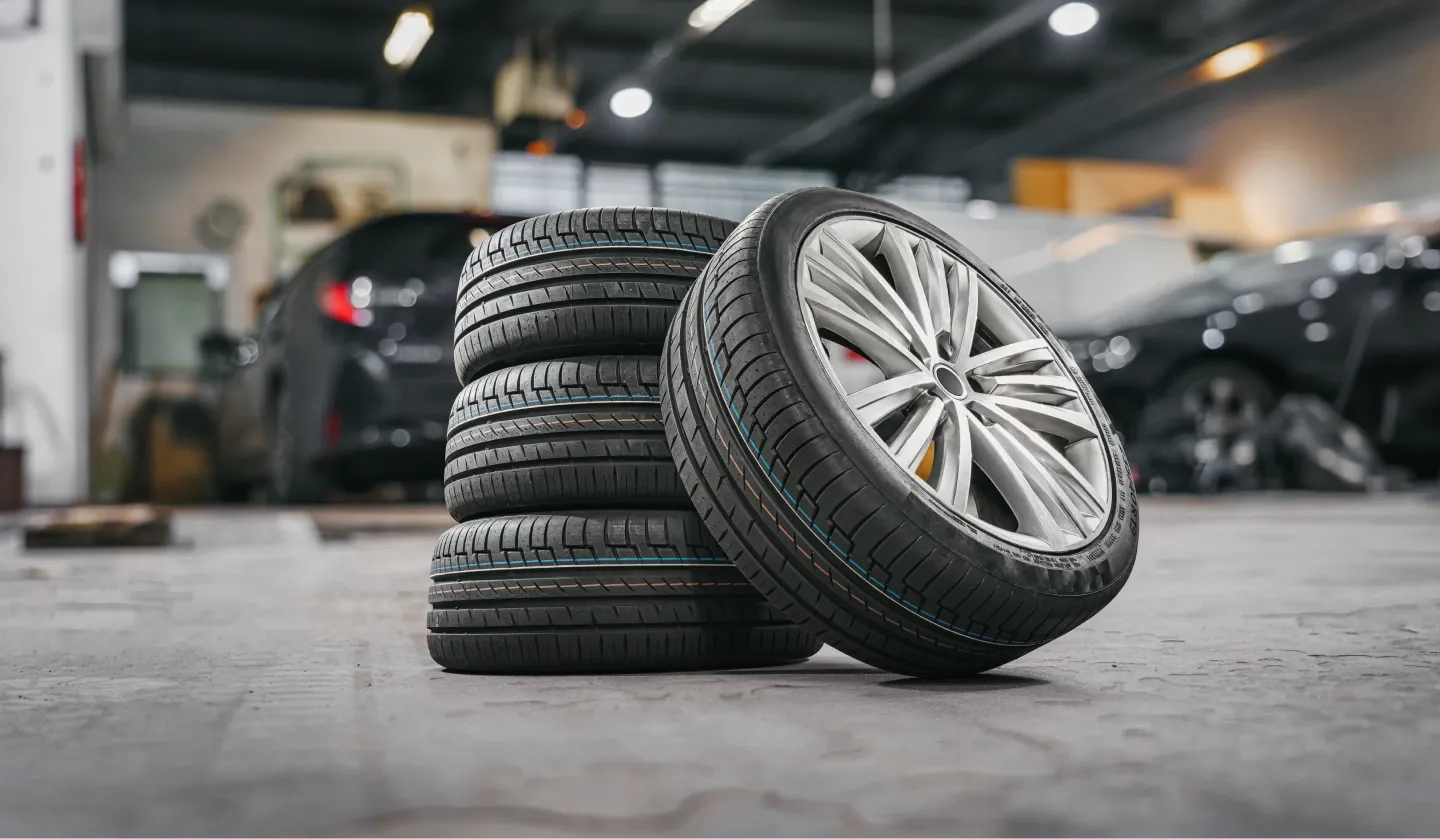 Four new car tires are stacked on a garage floor, with one tire standing upright and showing a silver alloy wheel. Blurred vehicles and workshop equipment are visible in the background.