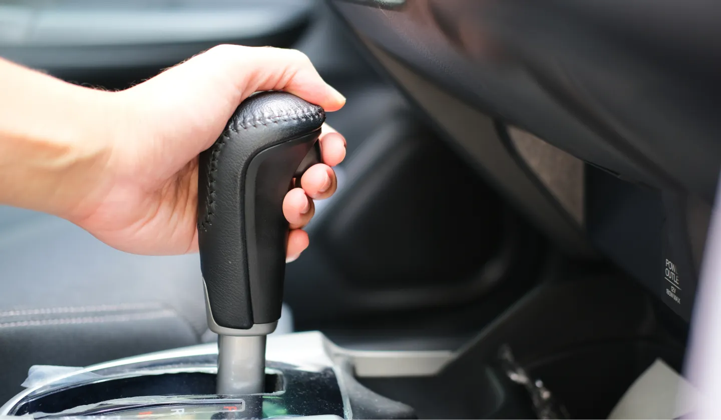 A close-up of a persons hand gripping an automatic car gear shift lever inside a vehicle. The interior details of the car, including the dashboard and center console, are partially visible.