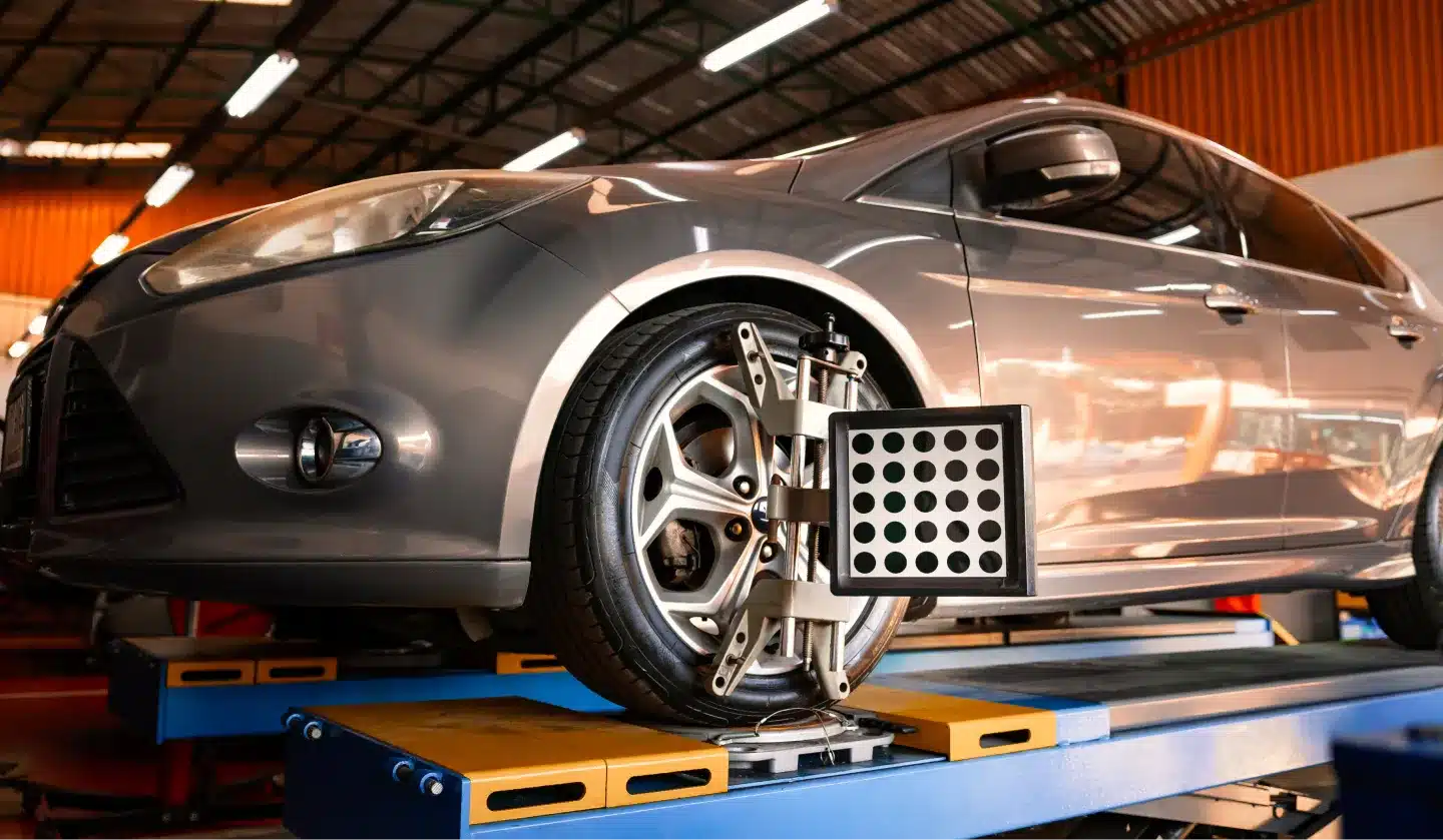 A gray car is elevated on a platform in an auto shop, with a wheel alignment tool attached to the front tire. The garage has orange walls and a metal roof.