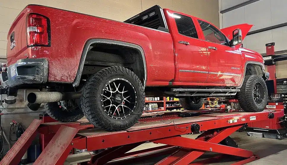 A red pickup truck with black wheels is parked on a hydraulic lift in an automotive workshop. The trucks hood is open, and various tools and equipment are visible in the background.